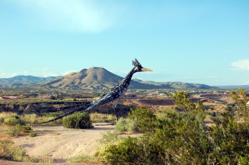 A large metal sculpture of a bird stands in a desert landscape with mountains in the background under a clear blue sky.