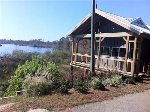 A cozy wooden house by a calm river, surrounded by greenery and colorful flowers under a clear blue sky.