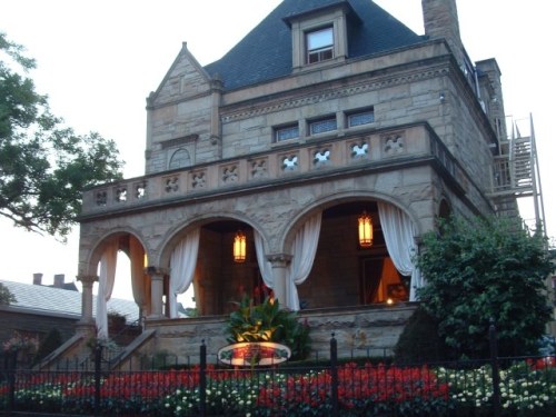 A historic stone mansion with a balcony, decorative lanterns, and vibrant flower beds in front.