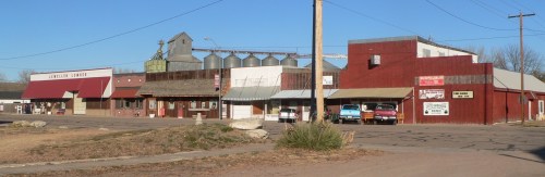 A small town street featuring several businesses, grain silos, and parked cars under a clear blue sky.