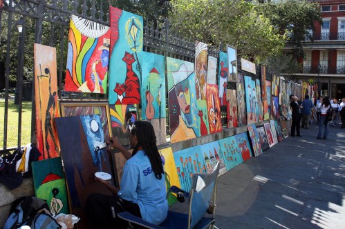 A street artist paints on a canvas, surrounded by colorful artwork displayed on a fence in a lively outdoor setting.