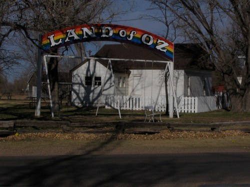 Colorful "Land of Oz" sign arching over a swing set in front of a white house, surrounded by trees and a picket fence.