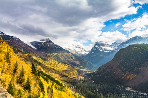 A scenic mountain landscape with autumn foliage, snow-capped peaks, and a winding river under a partly cloudy sky.