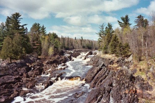 A rocky river flows through a forested landscape under a partly cloudy sky.