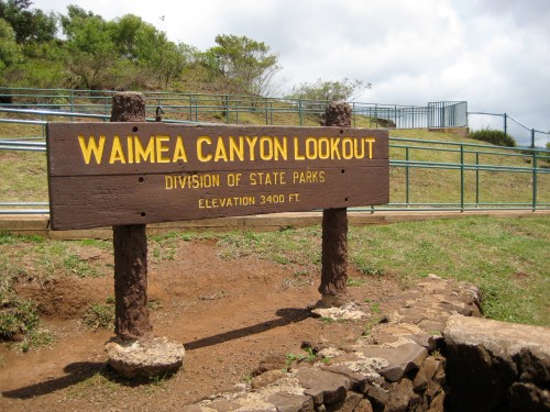 Sign for Waimea Canyon Lookout, Division of State Parks, elevation 3,400 ft, with a grassy area and cloudy sky in the background.
