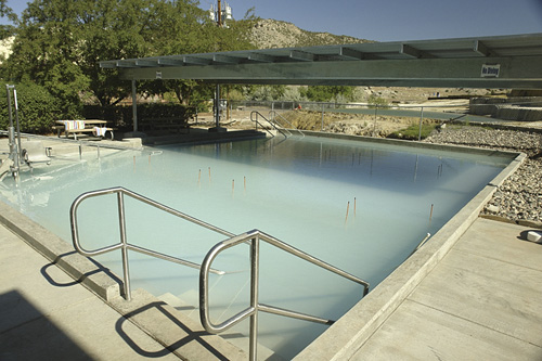 A clear swimming pool with metal railings, surrounded by a concrete deck and a shaded area, set against a mountainous backdrop.