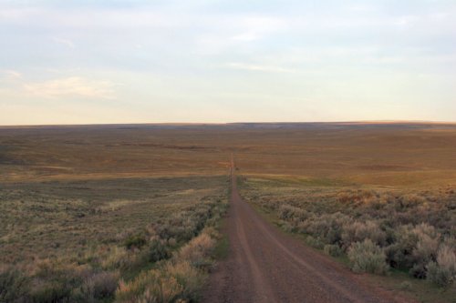 A long dirt road stretches through a vast, open landscape with sparse vegetation under a clear sky.