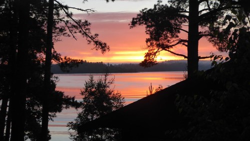 A serene sunset over a lake, framed by trees and a silhouette of a cabin in the foreground.