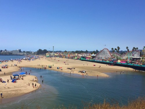 A sunny beach scene with people, colorful amusement rides, and palm trees under a clear blue sky.