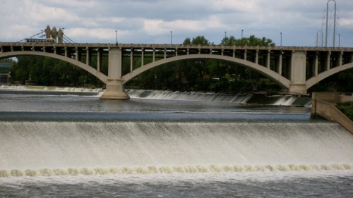 A bridge spans a river with a waterfall cascading beneath it, surrounded by greenery and a cloudy sky.