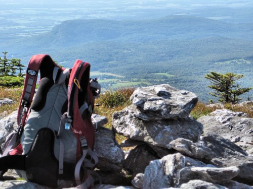 A backpack rests on a rocky summit, overlooking a vast green landscape and distant mountains under a clear sky.