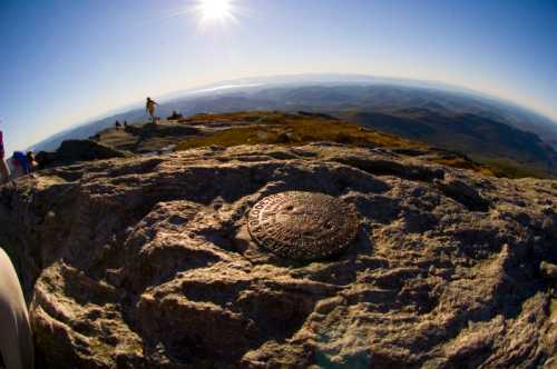 A sunlit mountain summit with a metal marker on a rock, overlooking a vast landscape and distant mountains.