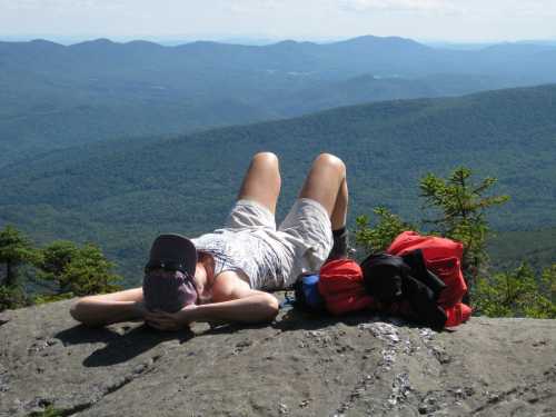 A person lies on a rock ledge, relaxing and enjoying a scenic mountain view with green hills in the background.