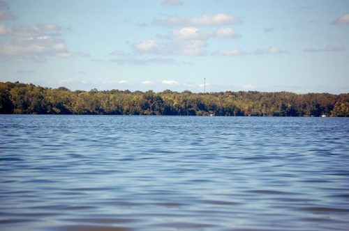 A calm lake with gentle ripples, surrounded by lush green trees under a partly cloudy sky.