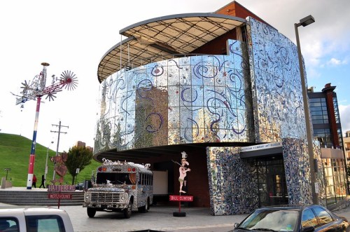 A colorful, mirrored building with a decorated bus in front, featuring artistic sculptures nearby.