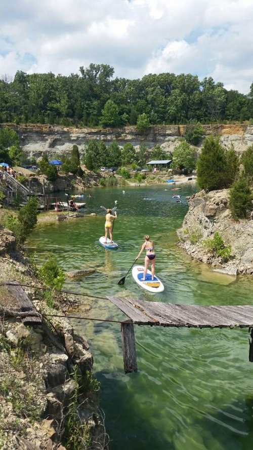 Two people paddleboarding on a green lake surrounded by rocky cliffs and trees on a sunny day.