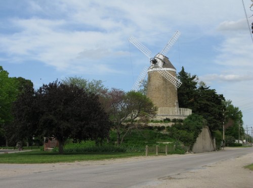 A historic windmill stands beside a road, surrounded by trees and greenery under a partly cloudy sky.