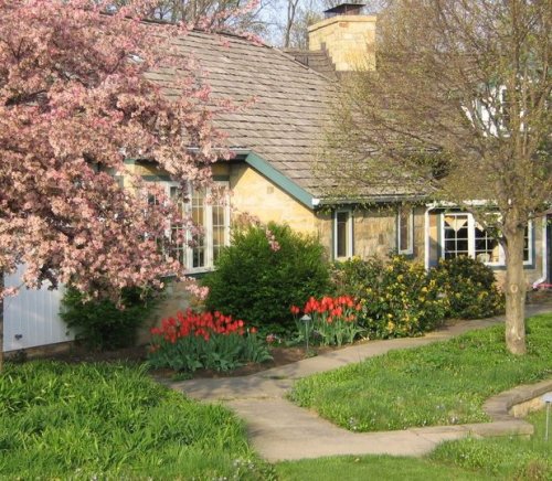 A charming house surrounded by blooming flowers and trees, with a winding path leading to the entrance.