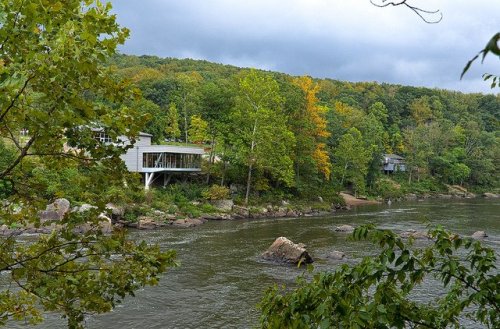 A serene river scene with modern houses nestled among lush green trees and autumn foliage along the bank.