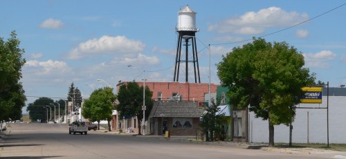 A small town street with a water tower in the background, lined with trees and buildings under a blue sky.