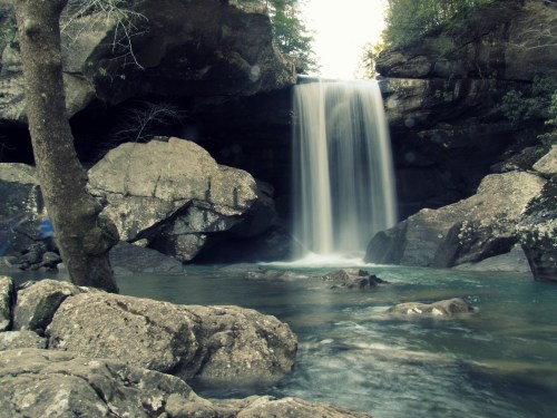 A serene waterfall cascades over rocks into a clear pool, surrounded by lush greenery and rugged terrain.
