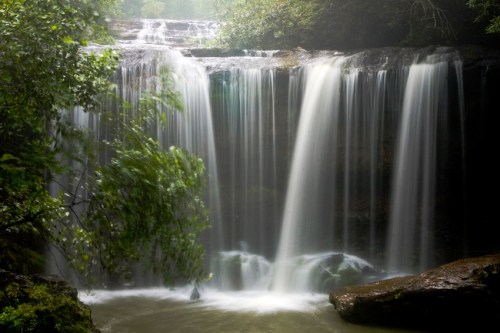 A serene waterfall cascading over rocks, surrounded by lush greenery and mist, creating a tranquil natural scene.