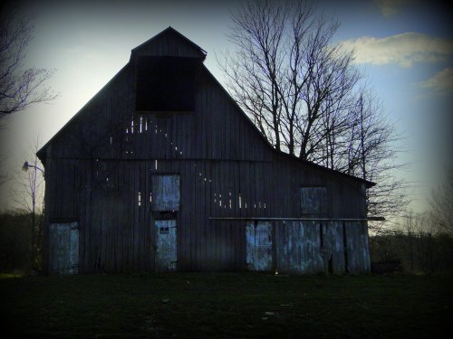 A weathered barn with a darkened upper window, surrounded by bare trees against a cloudy sky.