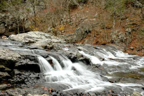 A serene waterfall cascades over dark rocks, surrounded by autumn foliage and rugged terrain.