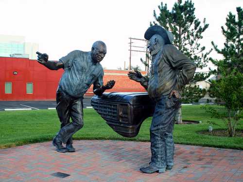 Two bronze statues of men conversing beside a classic car hood, set in a grassy area with trees in the background.