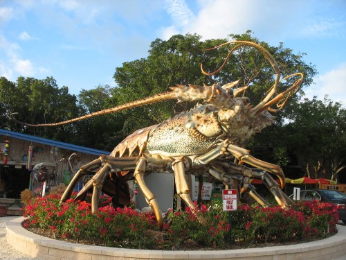 A large, colorful lobster sculpture surrounded by flowers, set against a blue sky and trees in the background.
