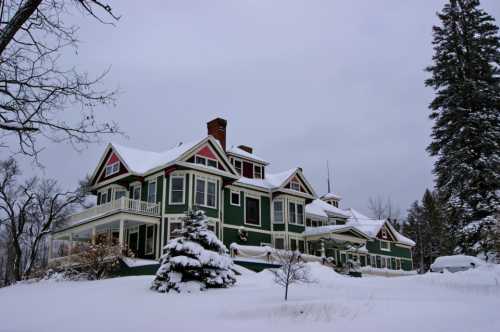 A large green house with red accents surrounded by snow-covered trees and a snowy landscape under a gray sky.