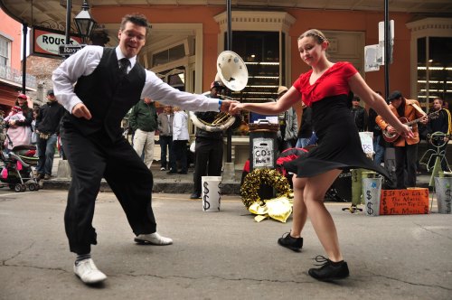 A man and woman dance joyfully on the street, surrounded by a lively crowd and musicians in the background.
