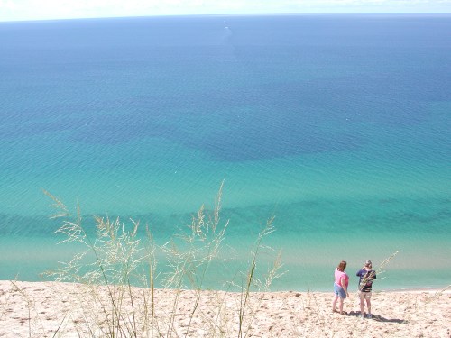 Two people stand on a sandy dune overlooking a calm, blue ocean with gentle waves and a clear sky.