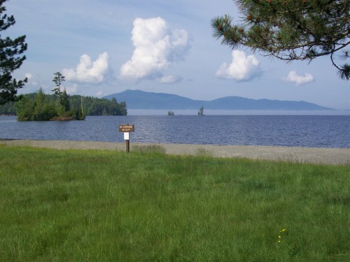 A serene lakeside view with grassy shore, distant mountains, and fluffy clouds in a blue sky. A sign is visible nearby.