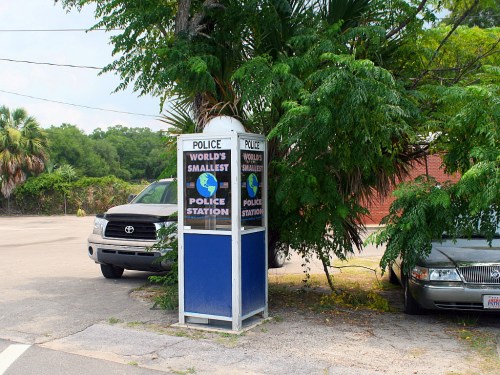 A small police station booth surrounded by greenery, with two parked cars nearby. Sign reads "World's Smallest Police Station."