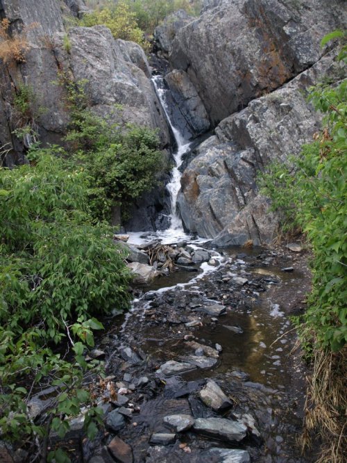 A small waterfall cascades over rocky terrain, surrounded by lush greenery and a tranquil stream below.