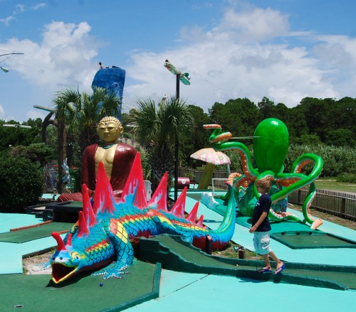 A colorful mini-golf course featuring a dragon, an octopus, and a statue, with a child walking nearby under a blue sky.