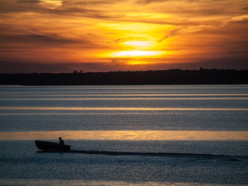 A lone boat glides across a calm lake at sunset, with vibrant orange and purple hues in the sky.