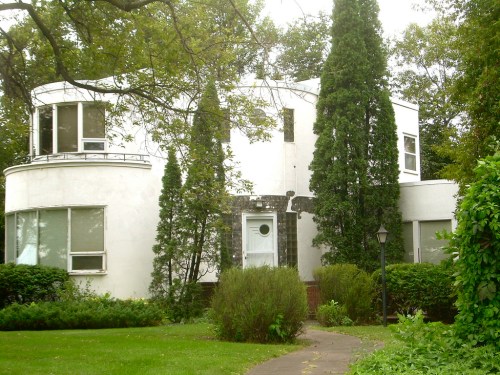 A modern white house with unique architecture, surrounded by greenery and tall trees.