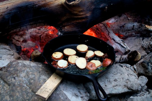 A cast iron skillet with sliced potatoes cooking over a campfire, surrounded by rocks and glowing embers.