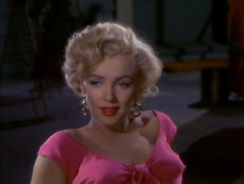 A glamorous woman with blonde curls wearing a pink top and large earrings, looking playfully at the camera.