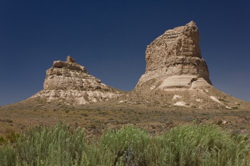 Two large rock formations rise from a grassy landscape under a clear blue sky.