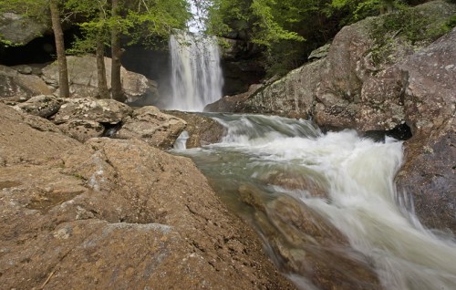 A serene waterfall cascades over rocks, surrounded by lush greenery and flowing water in a tranquil natural setting.