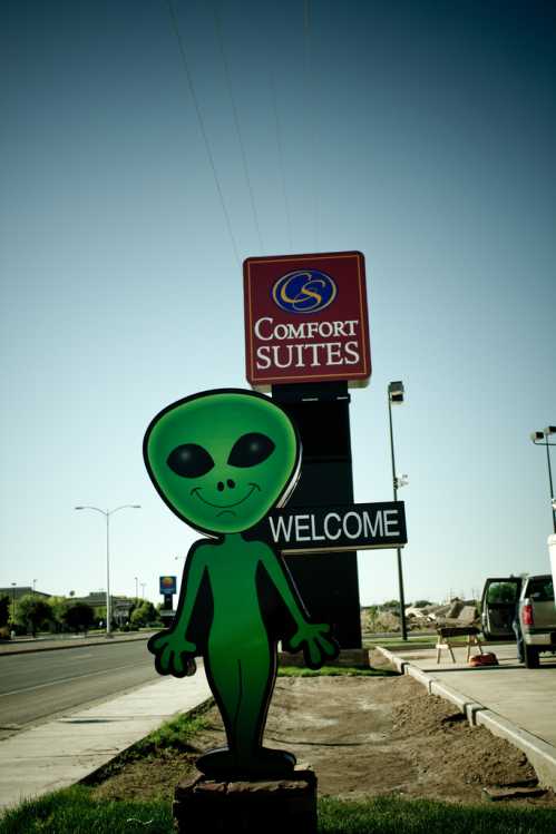 A green alien figure stands in front of a Comfort Suites sign, welcoming visitors. Clear blue sky in the background.