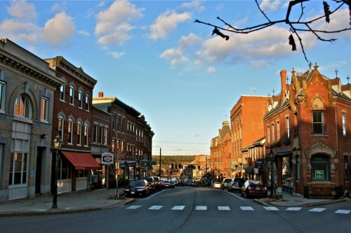 A charming small-town street lined with historic buildings, shops, and parked cars under a blue sky with scattered clouds.