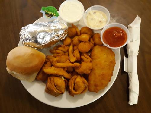 A plate of fried seafood, including shrimp, fish, and onion rings, with a baked potato, roll, and dipping sauces.