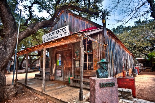 Historic wooden post office in Luckenbach, Texas, with a statue and trees surrounding the rustic building.