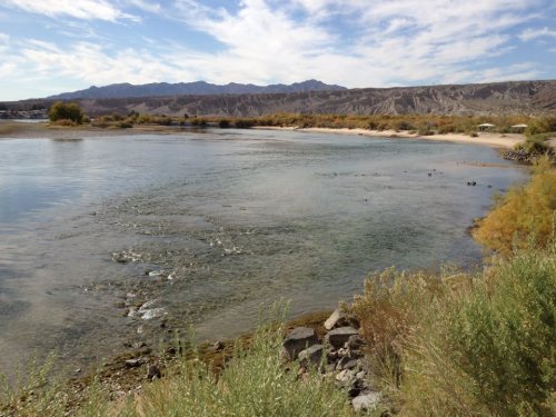 A serene river scene with gentle ripples, surrounded by sandy banks and distant mountains under a blue sky.