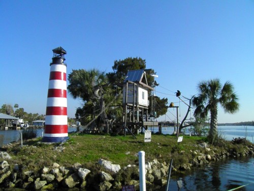 A colorful striped lighthouse beside a unique house on stilts, surrounded by water and palm trees under a clear blue sky.
