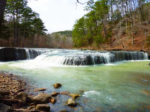 A serene waterfall cascades over rocky ledges into a clear, turquoise river, surrounded by lush green trees.